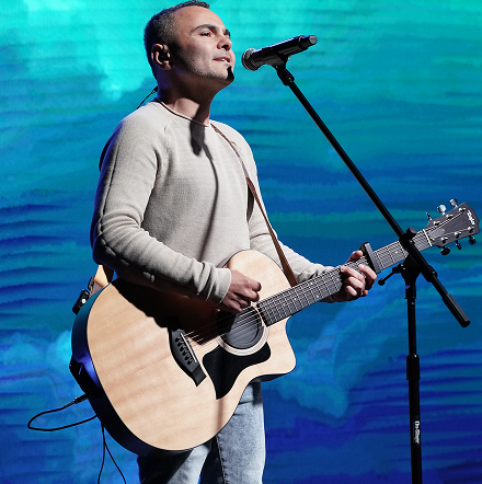 Singer playing acoustic guitar on stage, singing into a microphone against a blue backdrop.