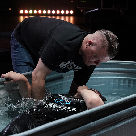 Man leaning over a water-filled tub, helping a person submerged in dark water under stage lights.
