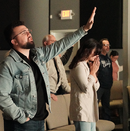 People standing in a room during worship, one man raising his hand and others with eyes closed.