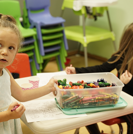 Children at a classroom table sharing a clear bin of colorful snacks and craft supplies