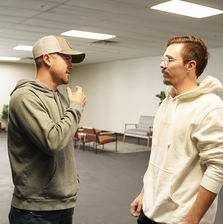 Two men talking in a bright office, one in a gray hoodie and cap, the other in a white hoodie.
