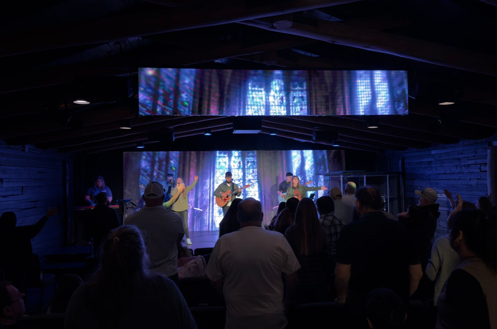 Dimly lit church stage with musicians performing before an audience, blue lights and stained-glass backdrop.