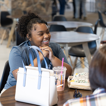 Smiling woman at a café table with a white handbag, iced drink, and sandwich