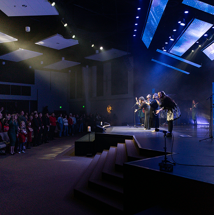 Musicians singing on a dimly lit stage in a church, with blue lights and an audience seated to the left.