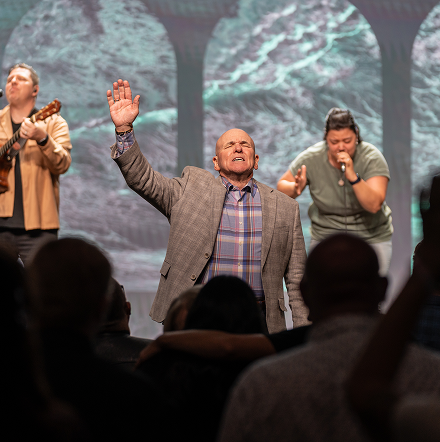 Three men on a stage with raised hands, singing before a blurred audience.
