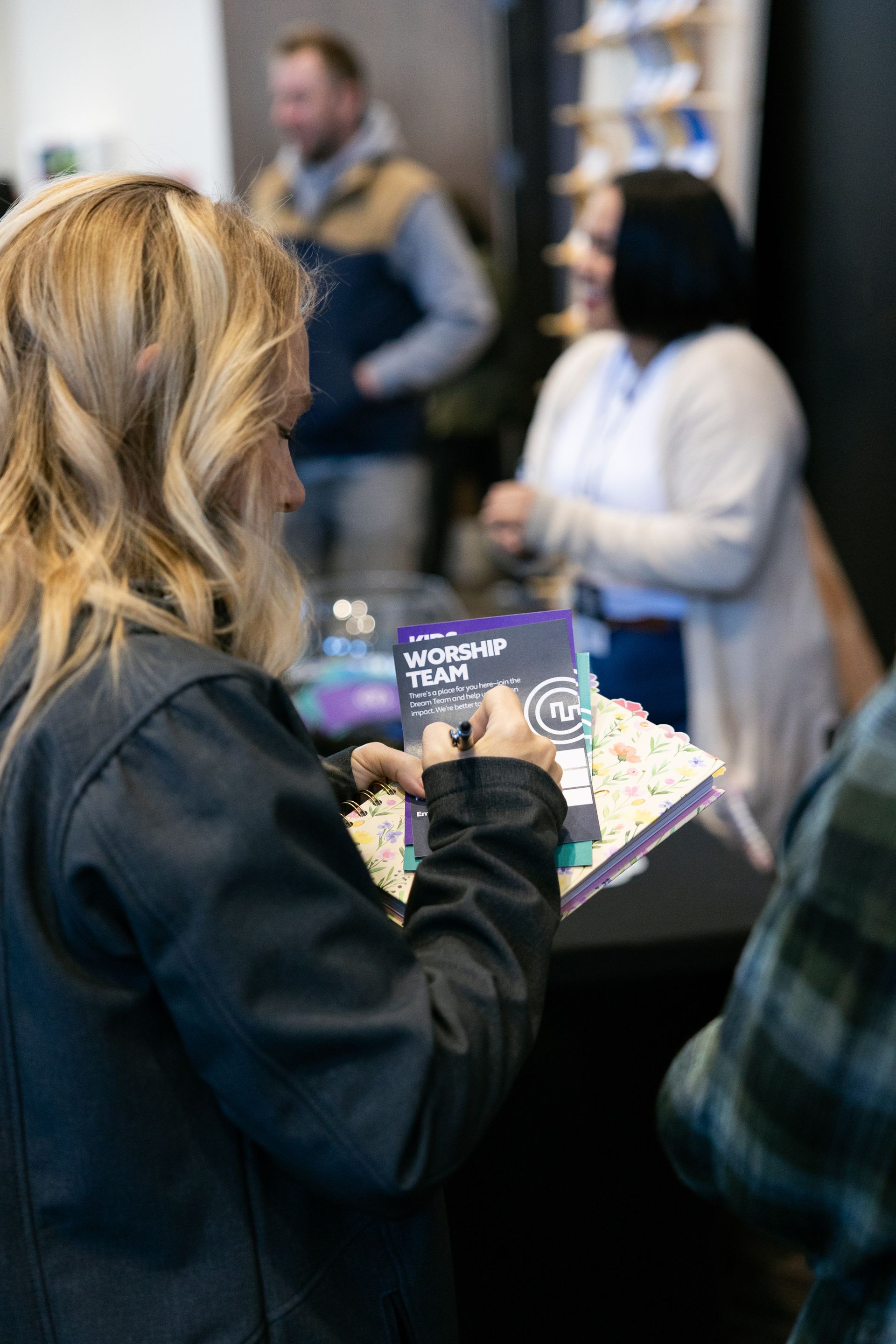 Person in black jacket holding a flyer in a crowded indoor event hallway.