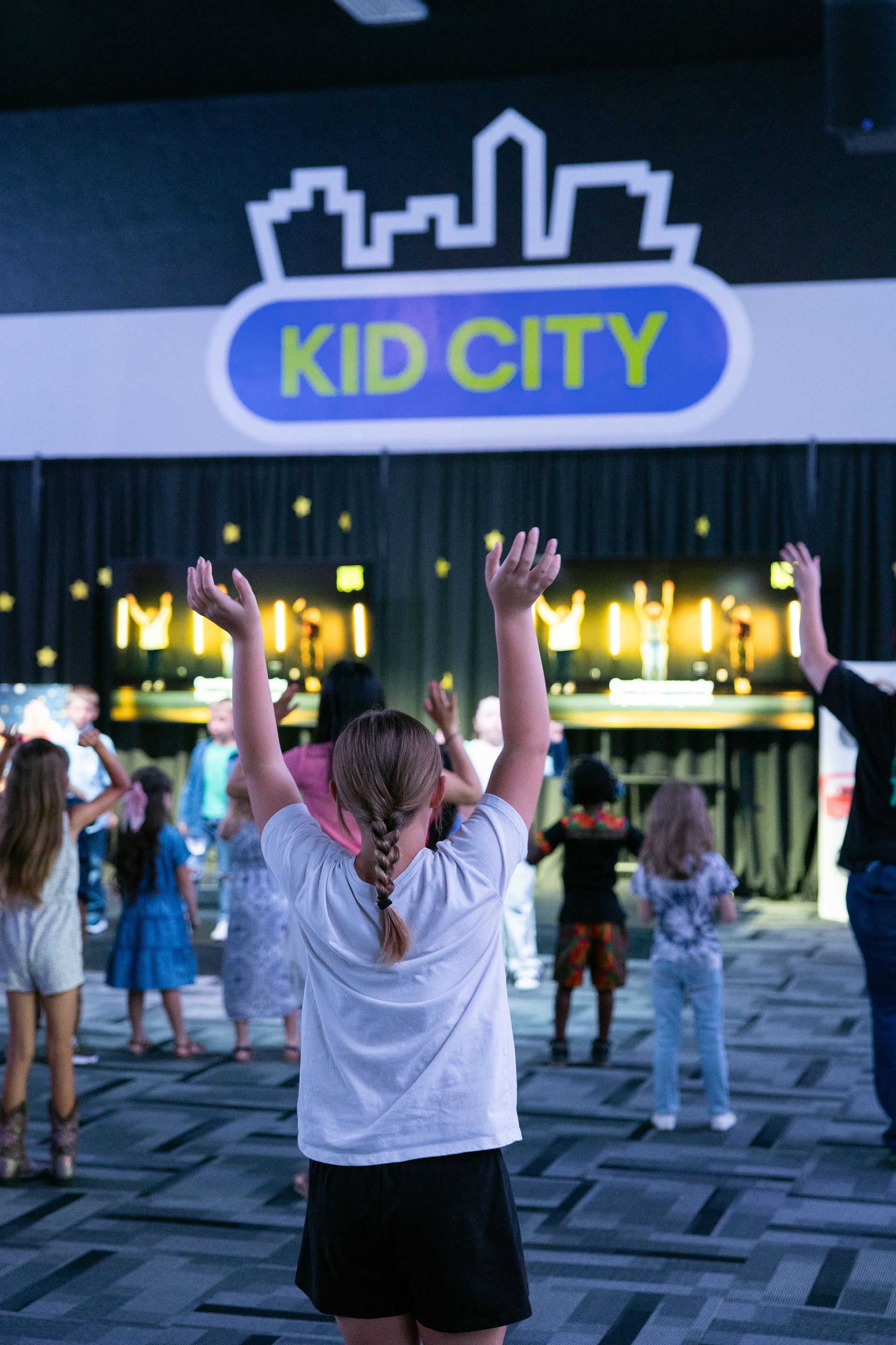 Children with raised arms in front of a lit “KID CITY” entrance at dusk