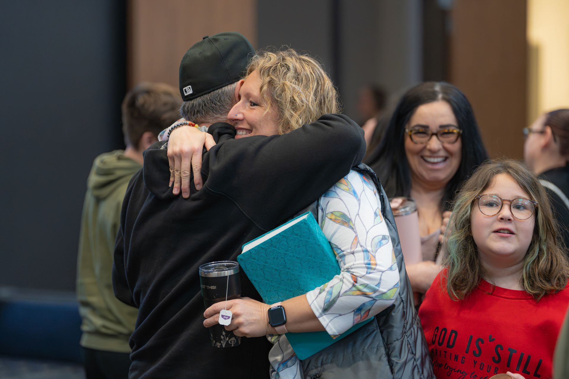 Two people hugging indoors, one holding a turquoise binder; others smile in the background.