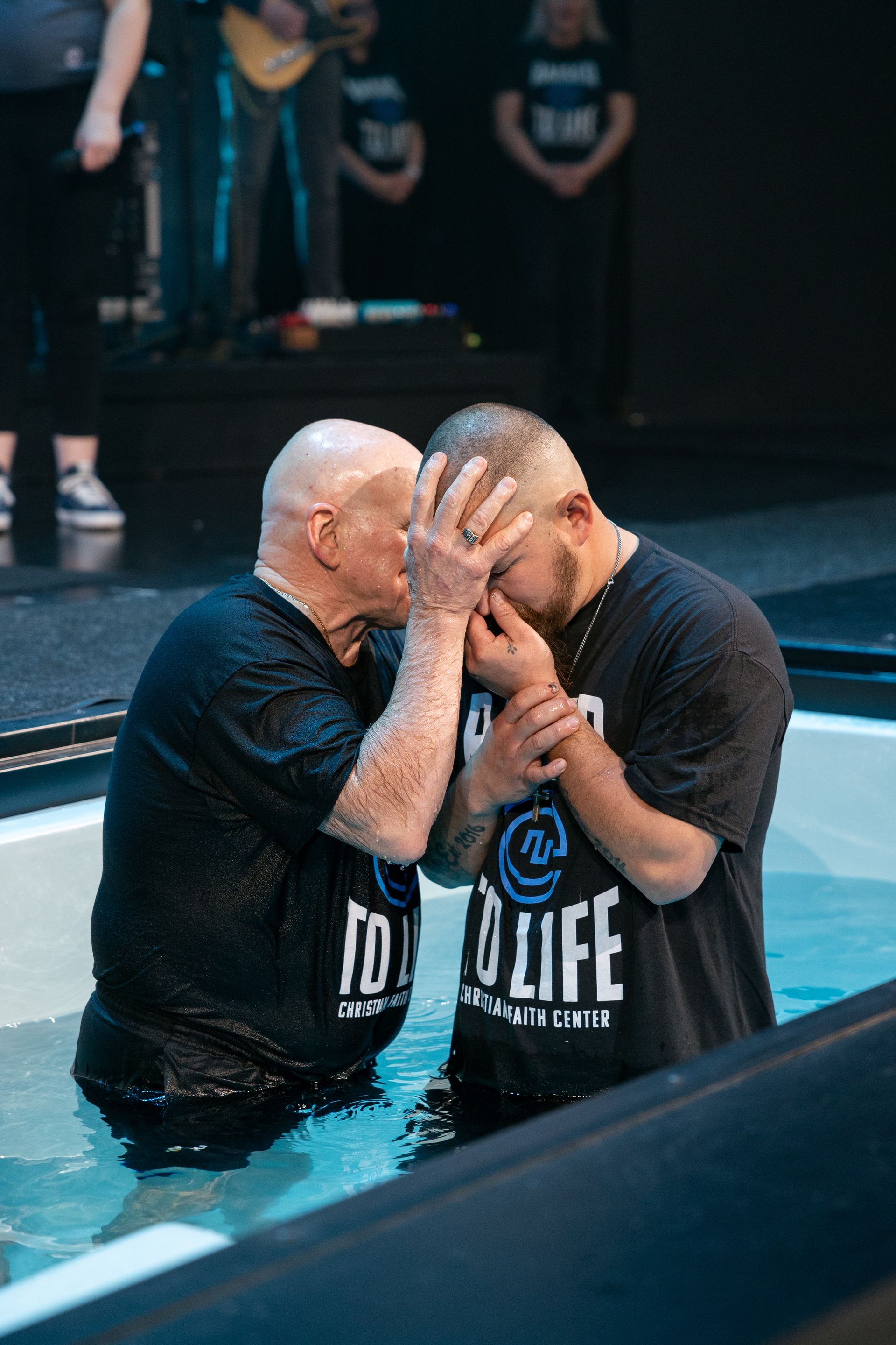 Two men in black shirts share an emotional embrace in a baptism pool.