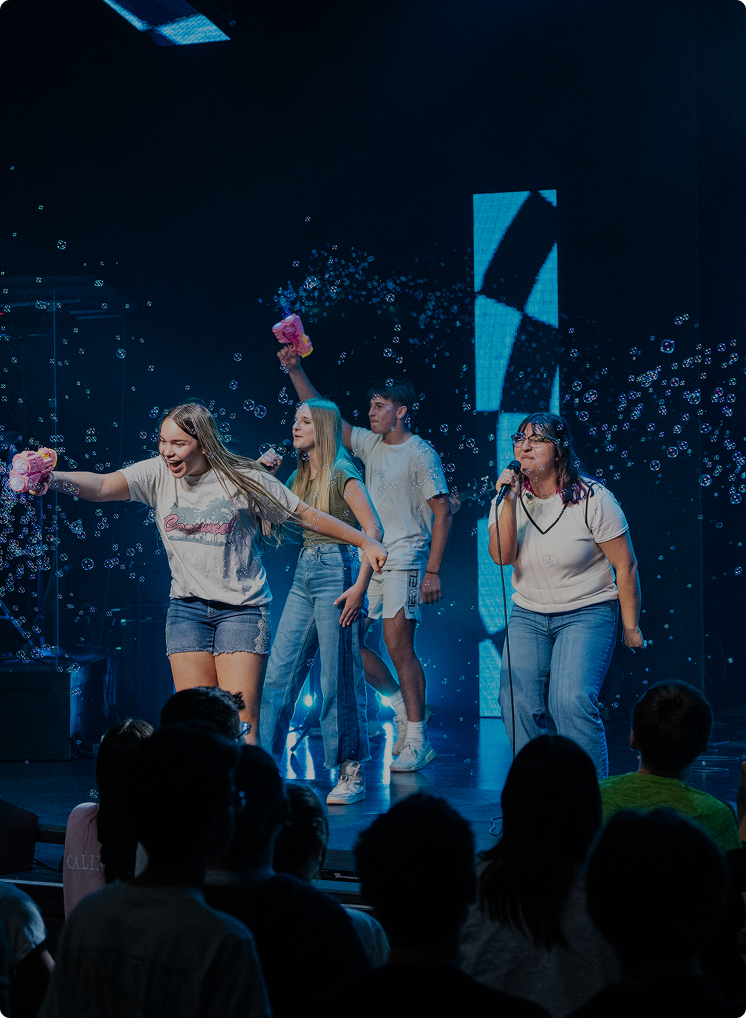 Four people onstage performing under blue lights with confetti, one holding a bouquet of flowers