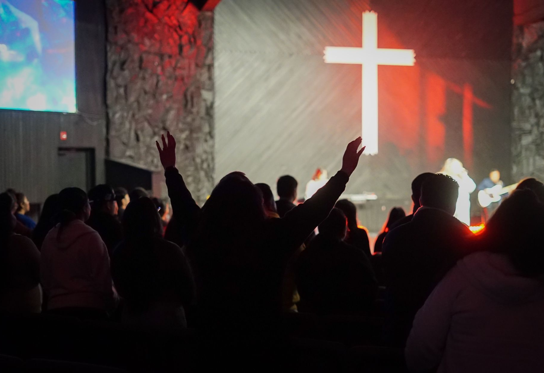 Silhouetted crowd with raised hands at a church service, facing a glowing red stage and illuminated cross.