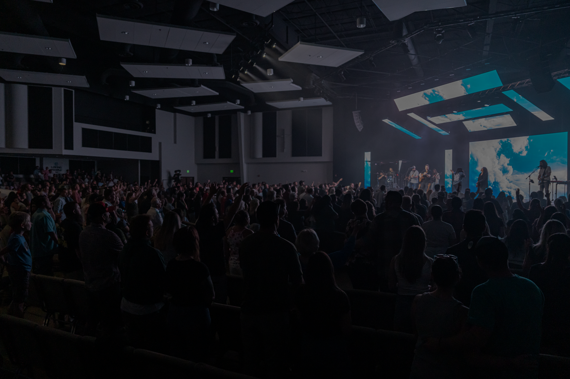 Crowded concert hall with a lit stage, blue lights, and a large audience in silhouette