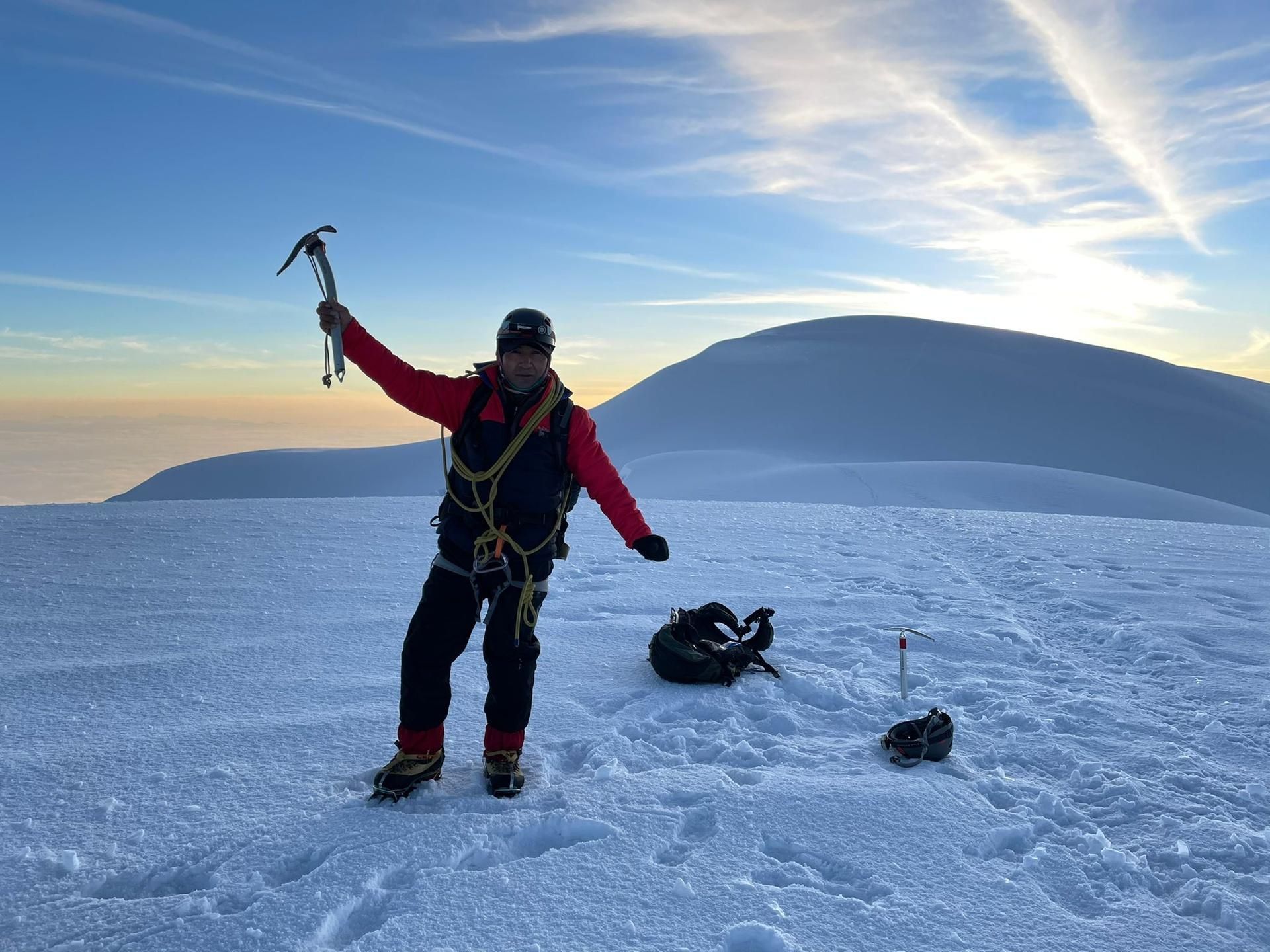 Mountaineer in red jacket stands on snowy ice, holding an ice axe with mountains and sea at sunset.