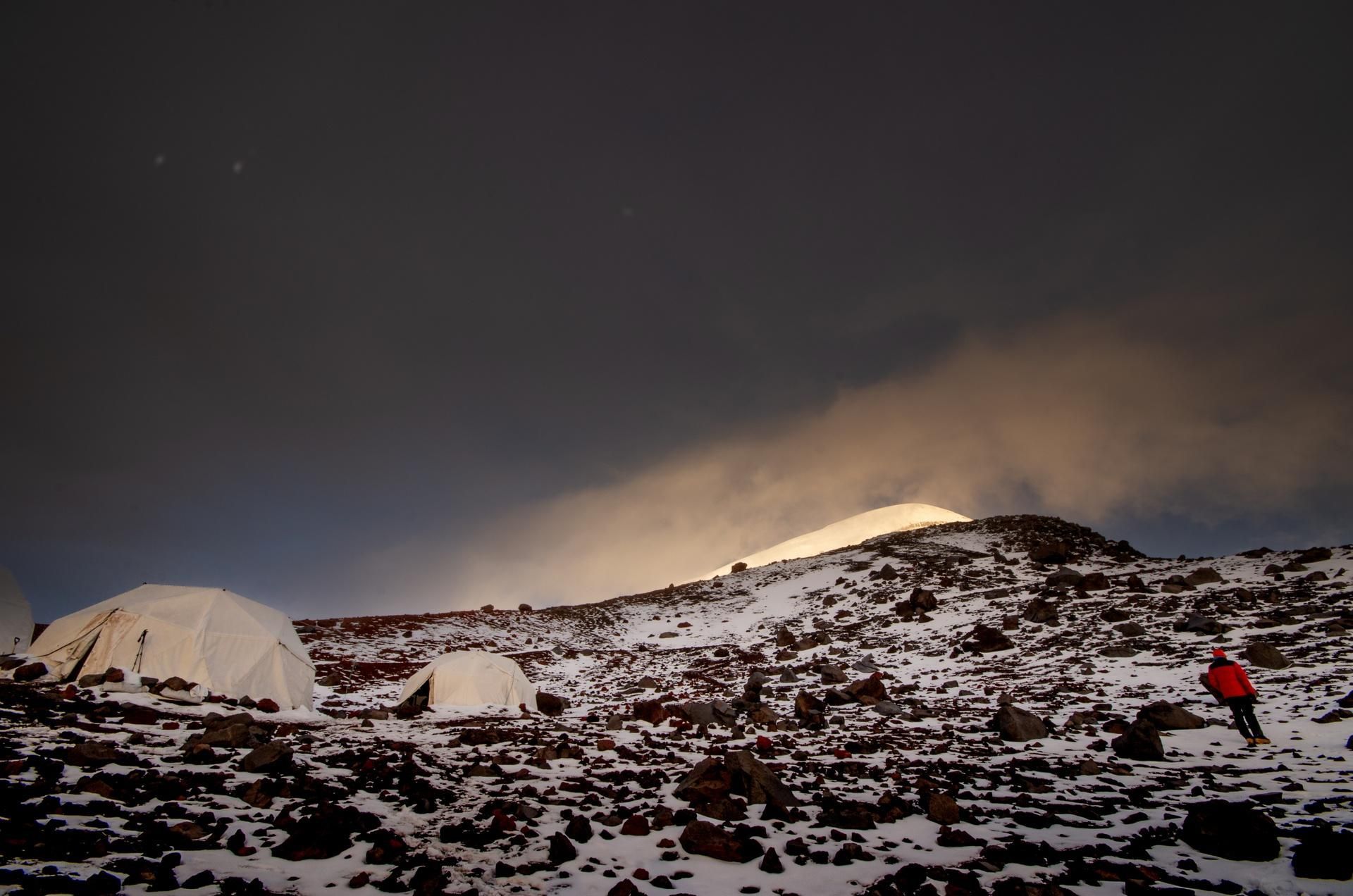 Snowy mountain campsite at night with tents, rocky ground, and a person in a red jacket on the right.