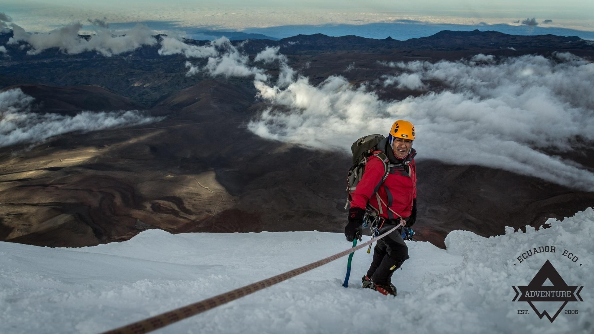 Climber in red jacket and orange helmet ascending snowy peak with rope, overlooking a volcanic landscape and clouds