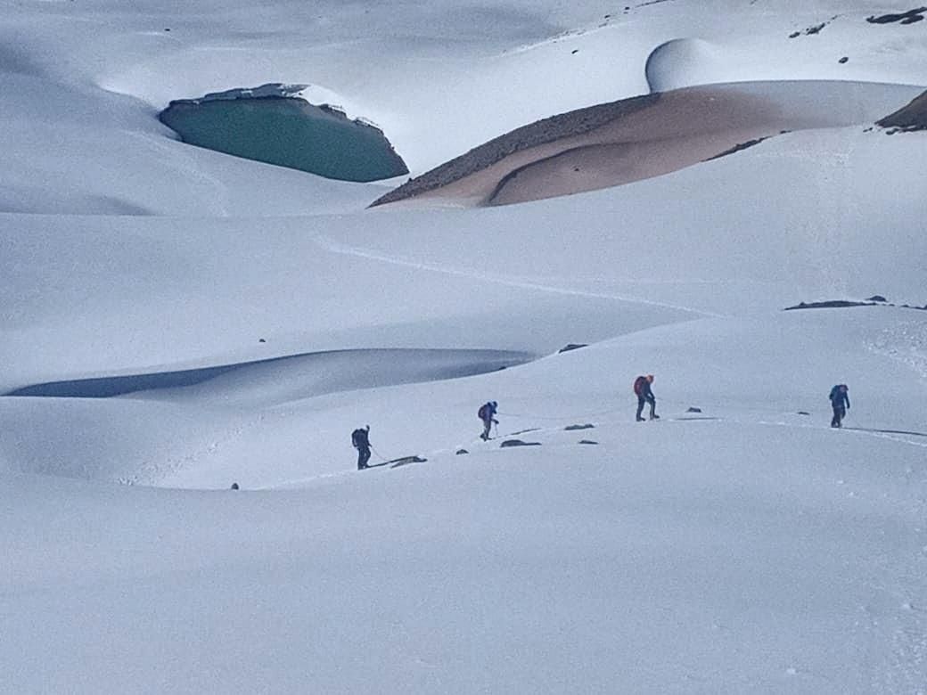 Skiers crossing a snowy mountain landscape with a turquoise glacial pool in the background