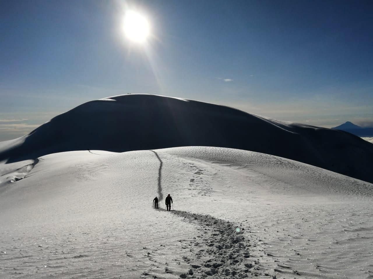 Two hikers on a snowy mountain ridge under bright sun, with footprints leading up the slope