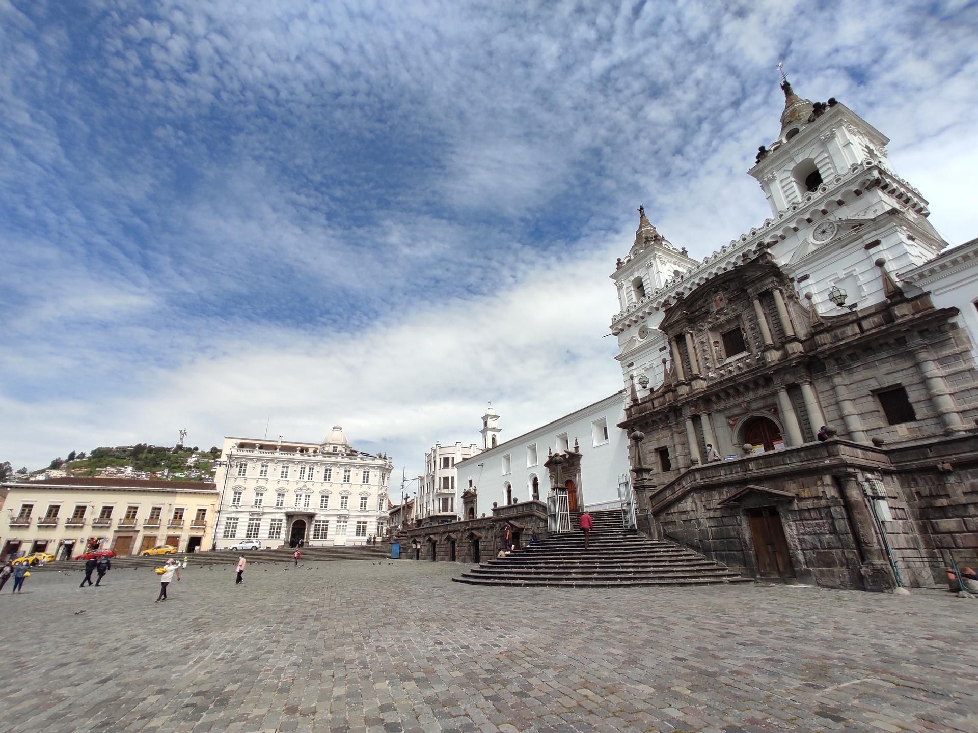 Historic white church on a cobblestone plaza under a cloudy blue sky, with a few people in the distance.