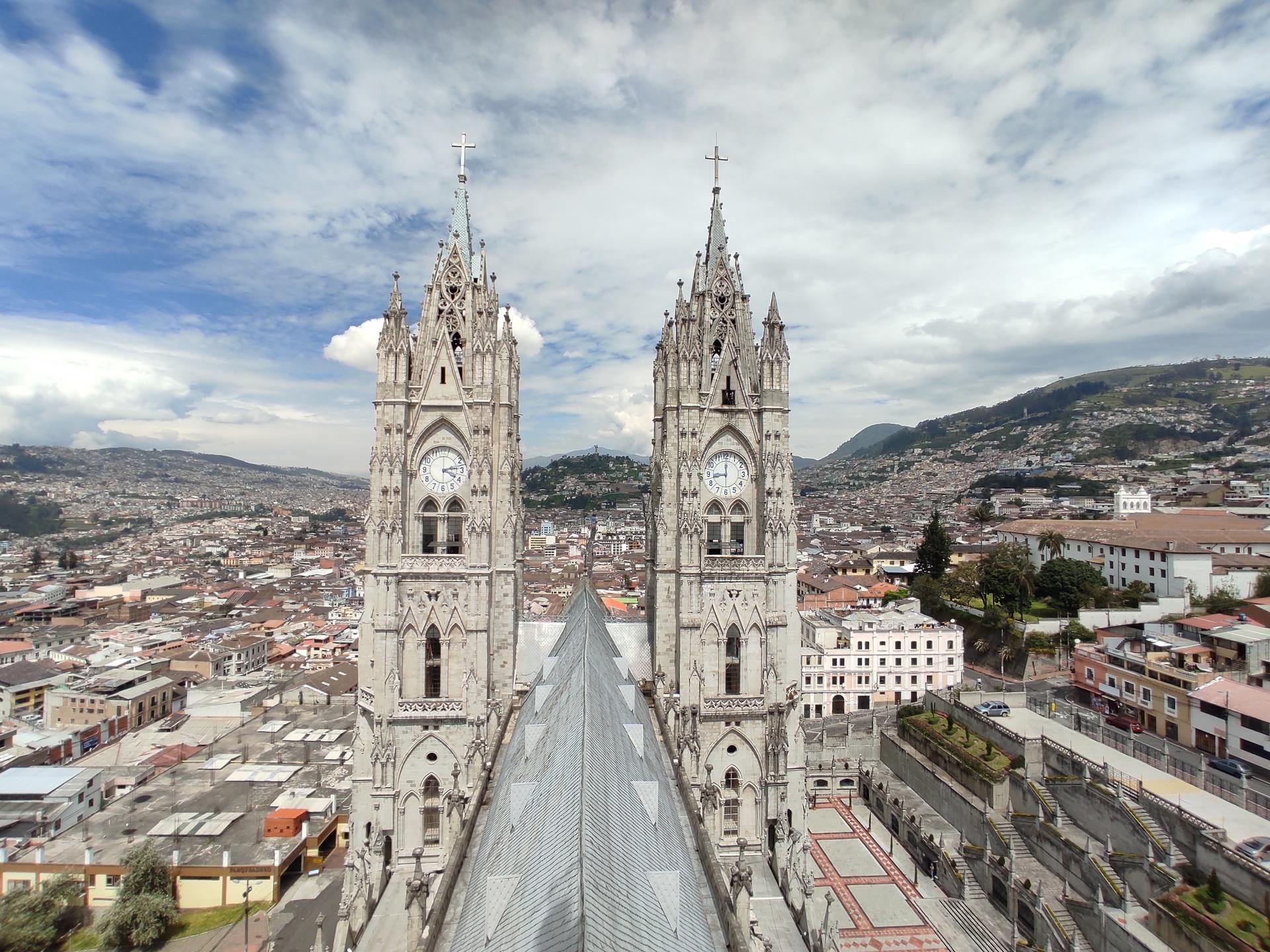 Aerial view of a cathedral with twin spires overlooking a dense city and distant hills under a cloudy sky
