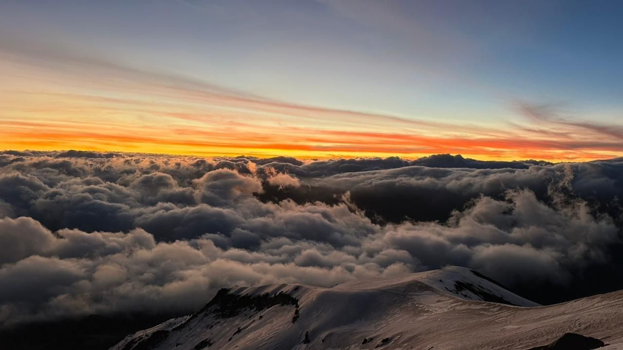Sunset above a sea of clouds, with orange and pink light over snow-covered peaks