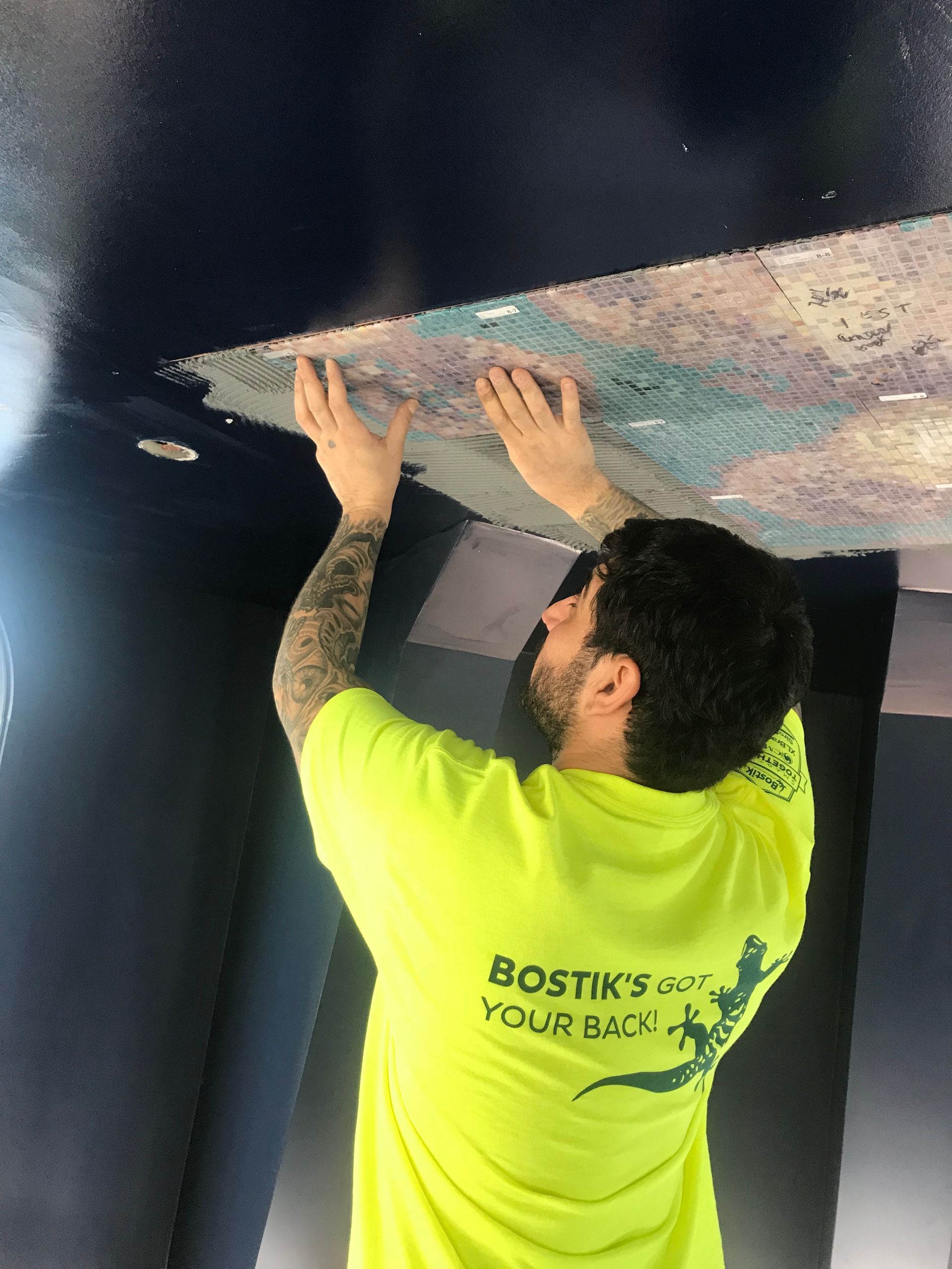 Man in yellow shirt installs ceiling tile. Dark blue walls, patterned tile.