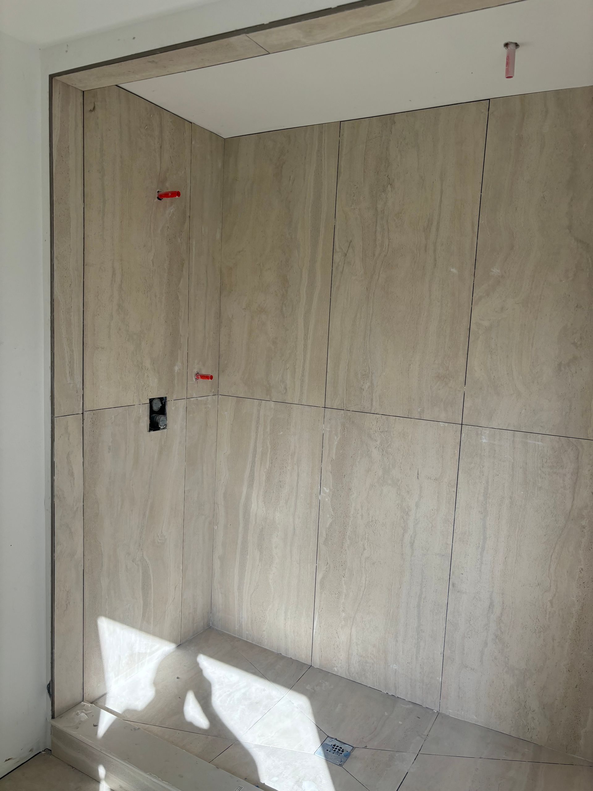 Shower stall under construction, beige tile walls with marked lines, unfinished ceiling and floor, plumbing visible.