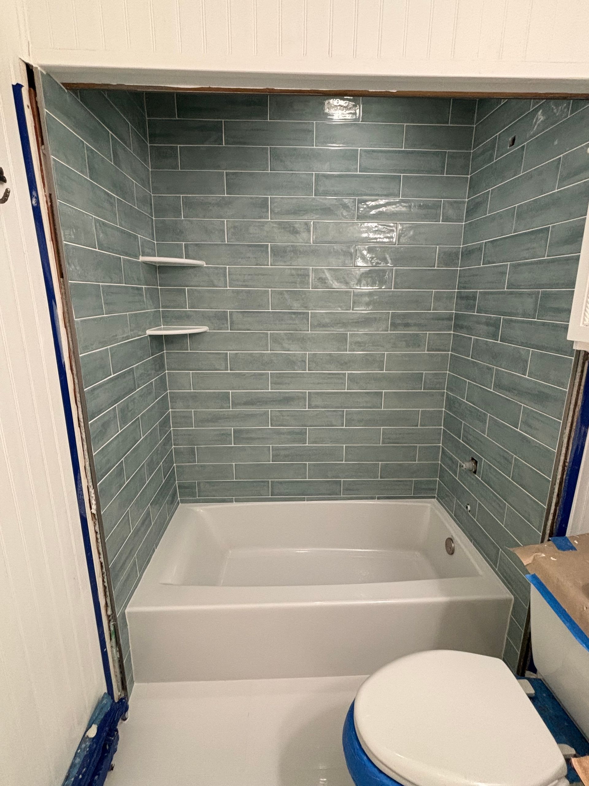 Bathroom with blue-tiled shower walls, white tub, shelves, and toilet.