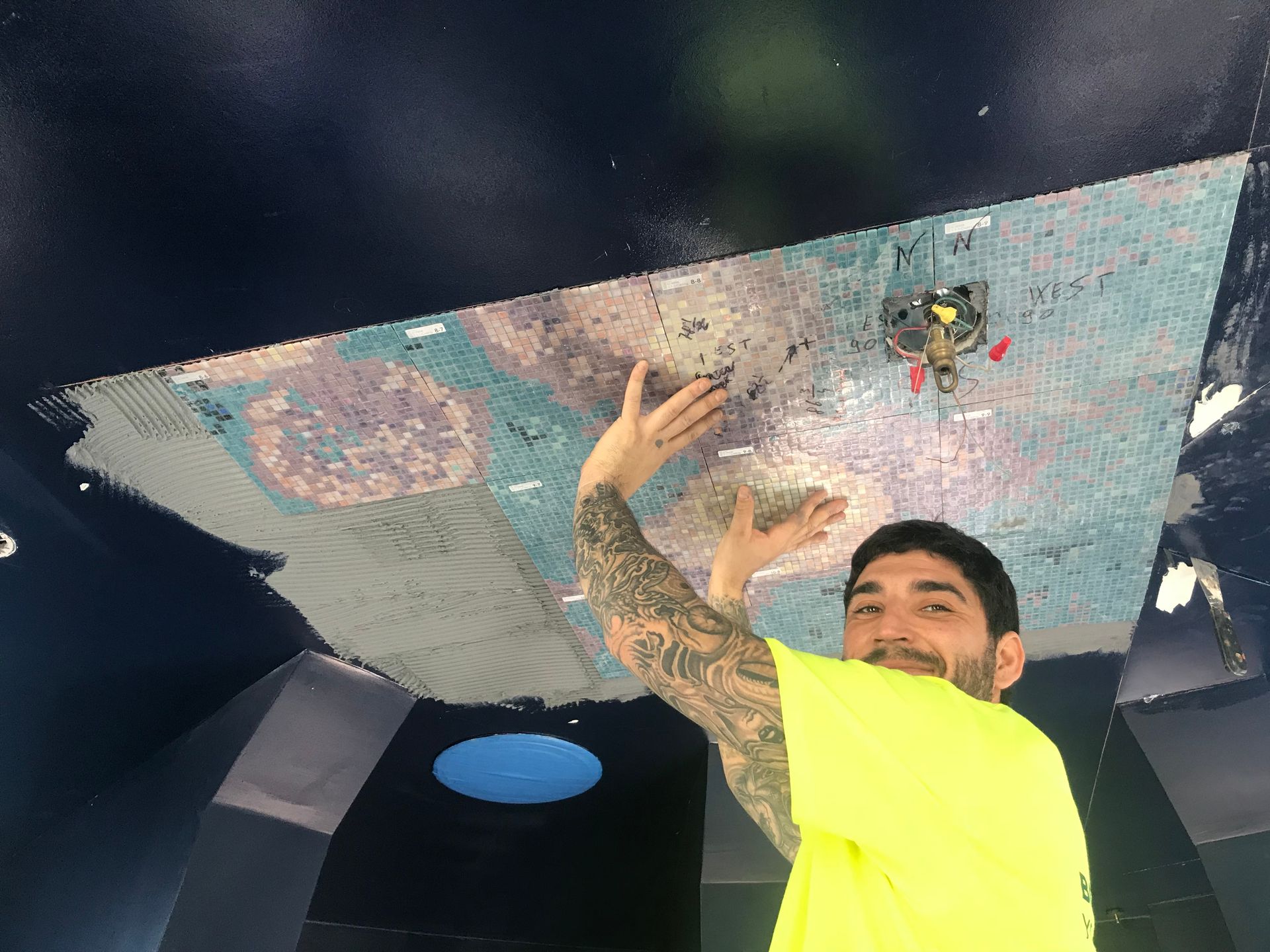 Man in neon yellow shirt, tattoos, smiling, holding up a large ceiling panel with colorful designs in a dark blue room.