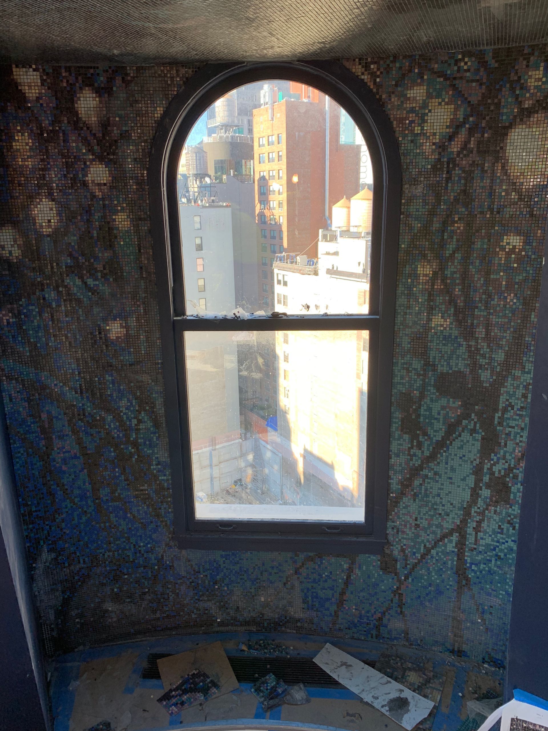 Arched window overlooking city buildings, surrounded by blue mosaic wall with spiderweb design.