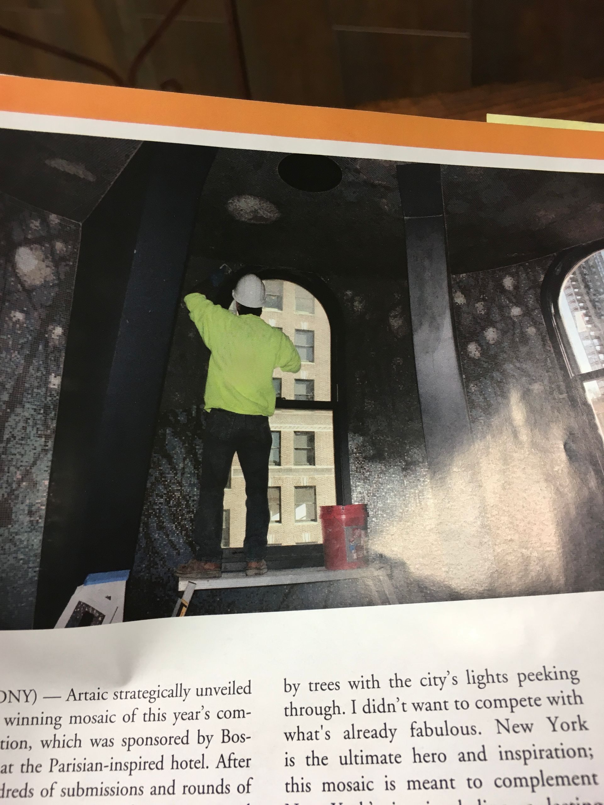 Person in a neon shirt painting a dark-colored interior space, seen through a window with a city view.