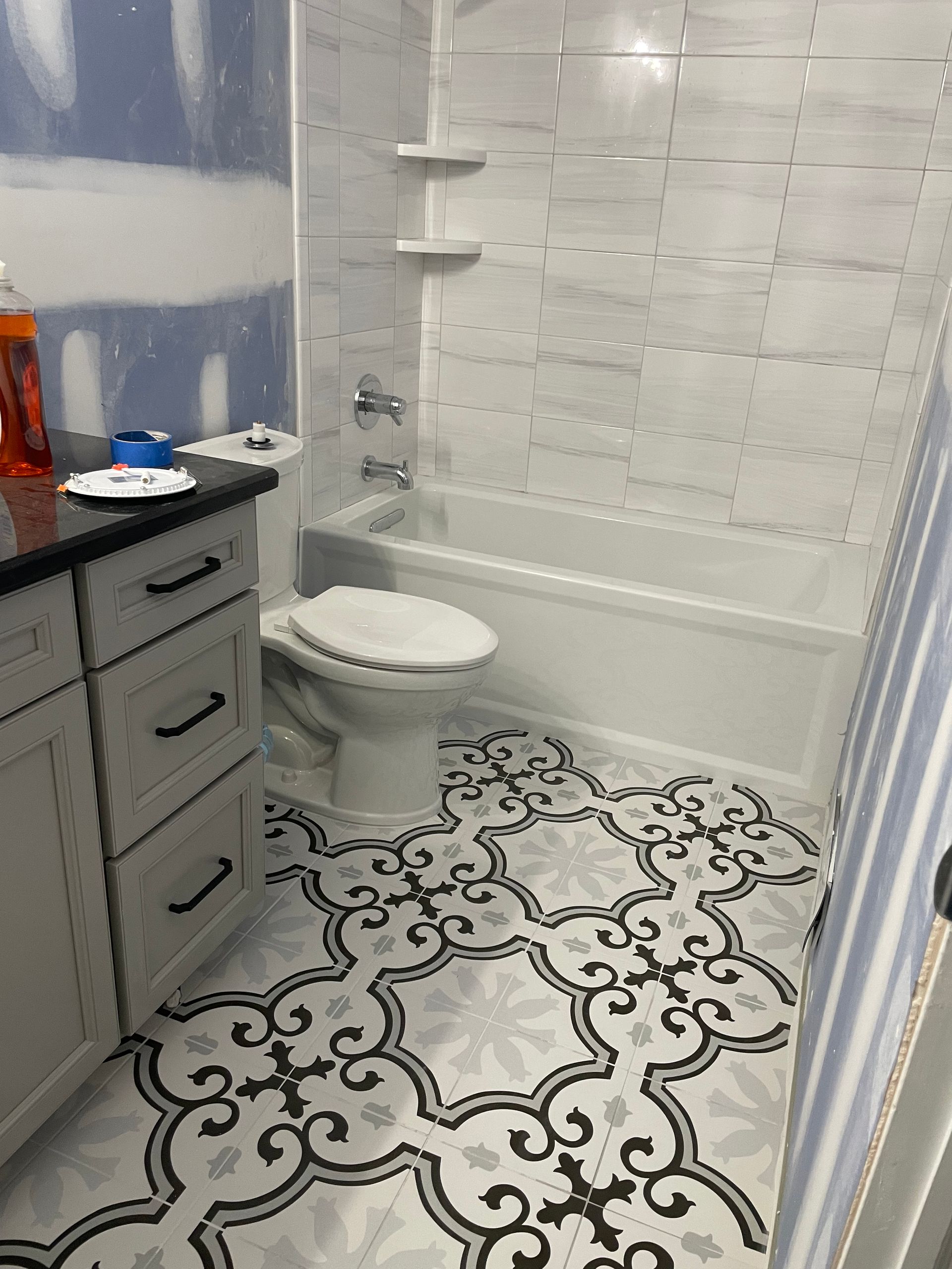 Bathroom with white bathtub, toilet, and patterned floor. White tile walls with shelves. Gray vanity.