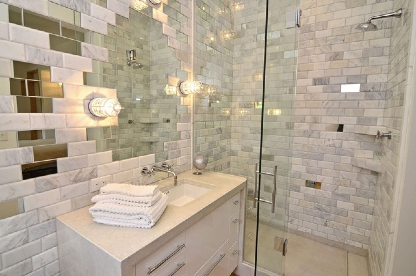 Bathroom with white and mirrored tile, glass shower, and white vanity.