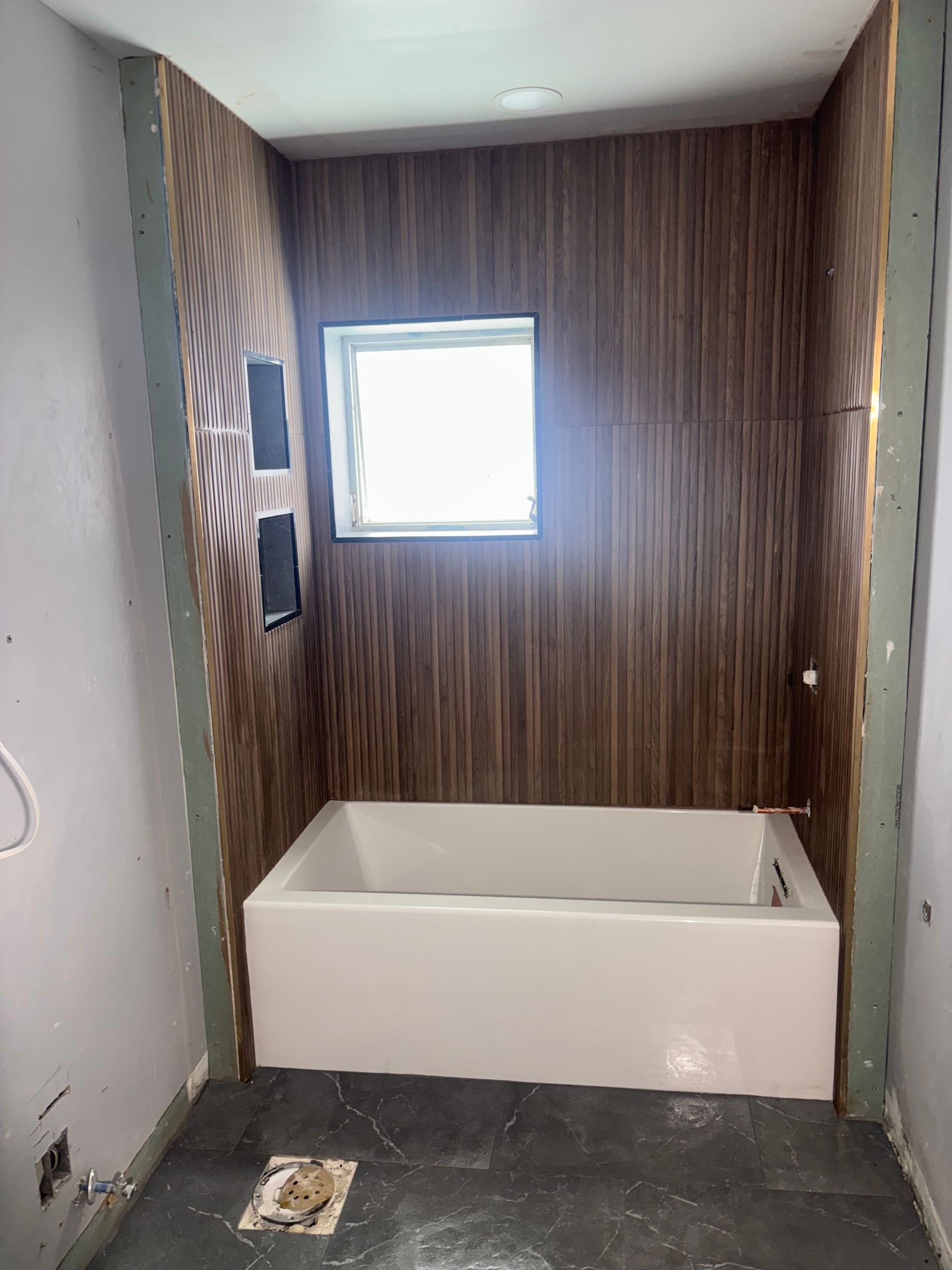 A bathroom under construction with a white tub, wood-paneled wall, small window, and unfinished walls.