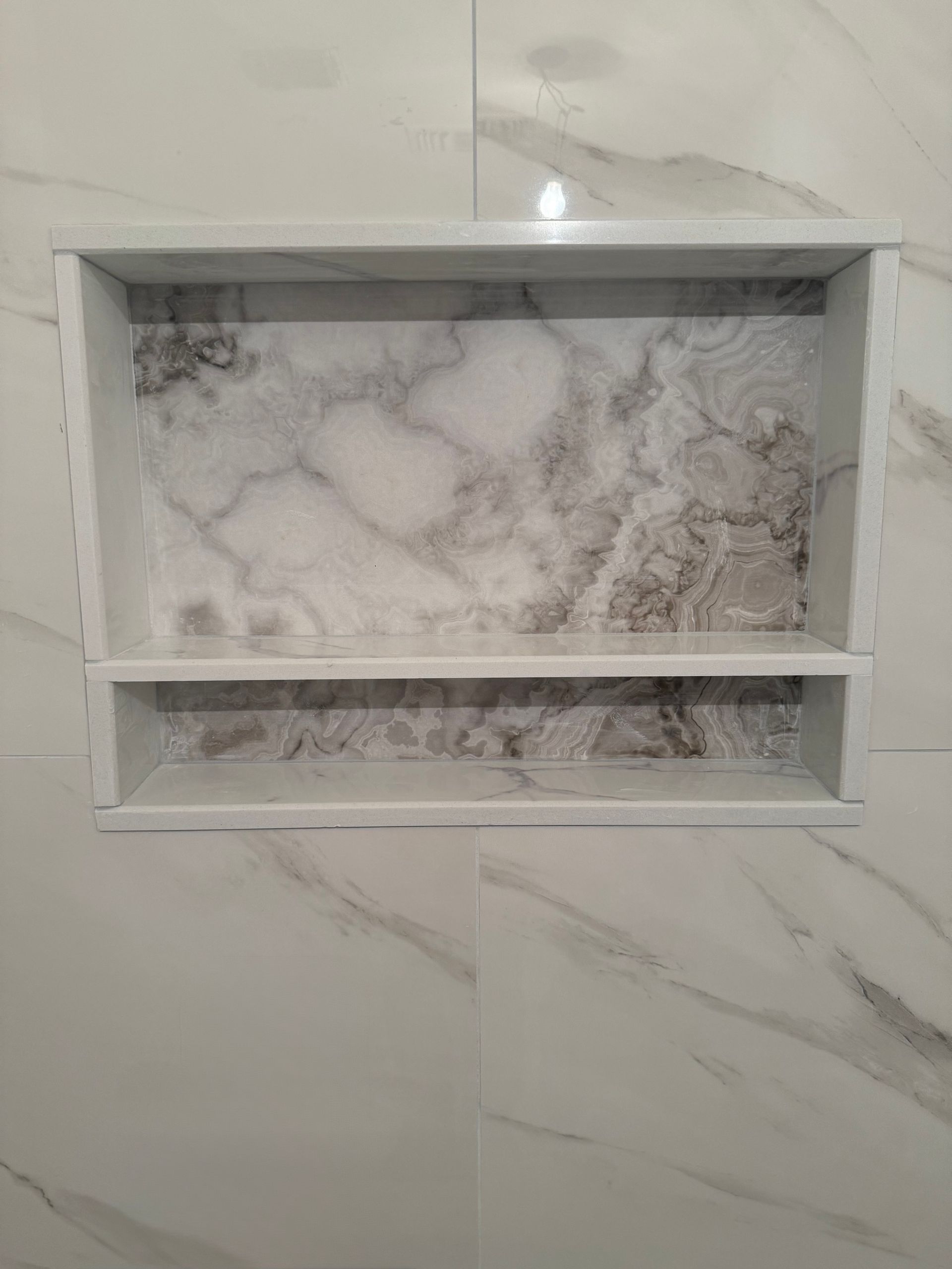 Recessed rectangular shower shelf with marble-patterned backing and a white shelf. It is set into white tiled wall.
