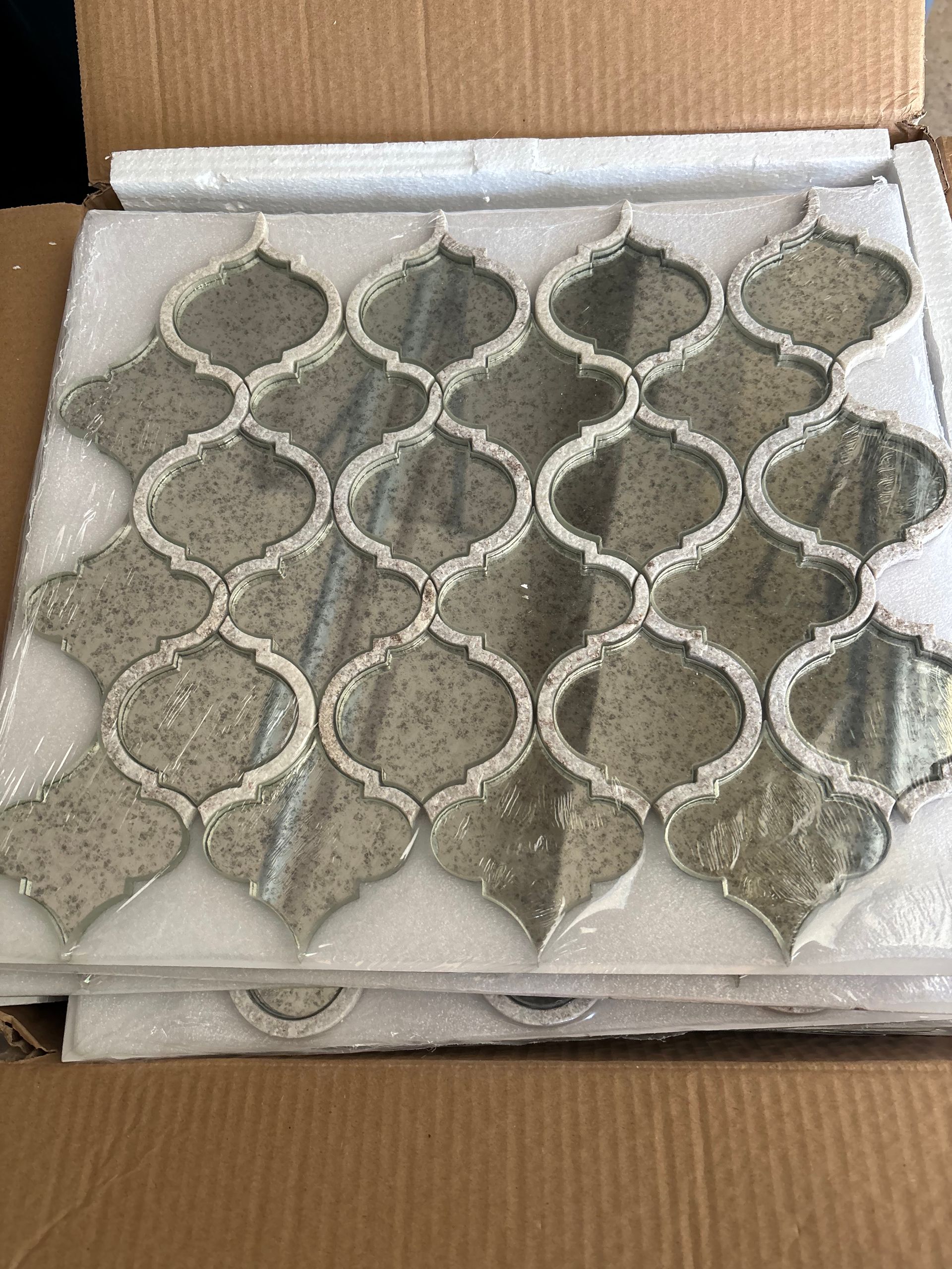 Arabesque-shaped glass mosaic tiles in a box. Gray and gold colors.