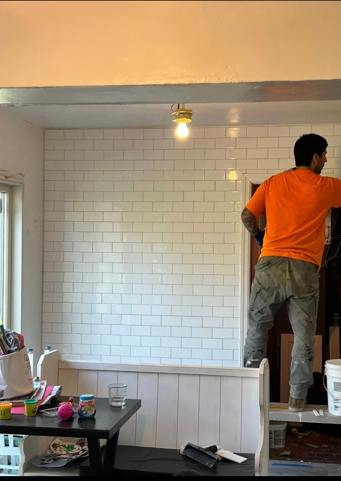 Person in orange shirt painting white subway tile wall.