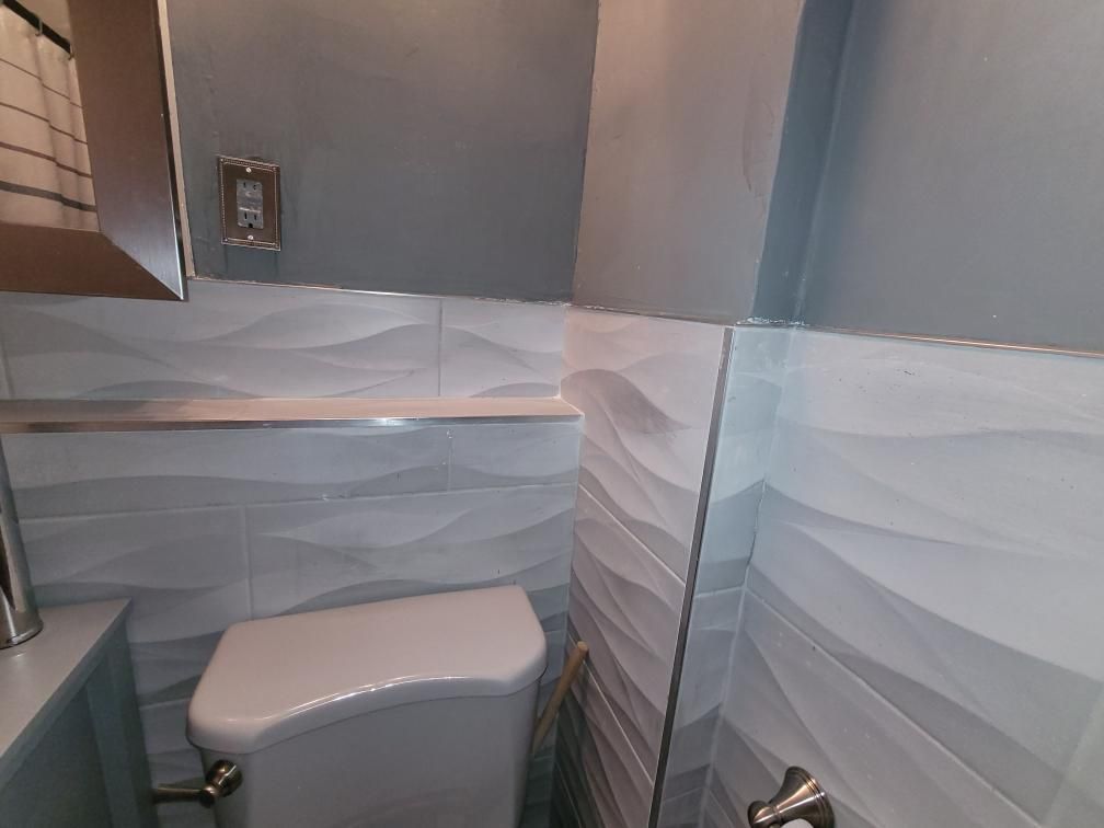 Bathroom corner with patterned white and blue wave tiles, toilet, and gray walls.