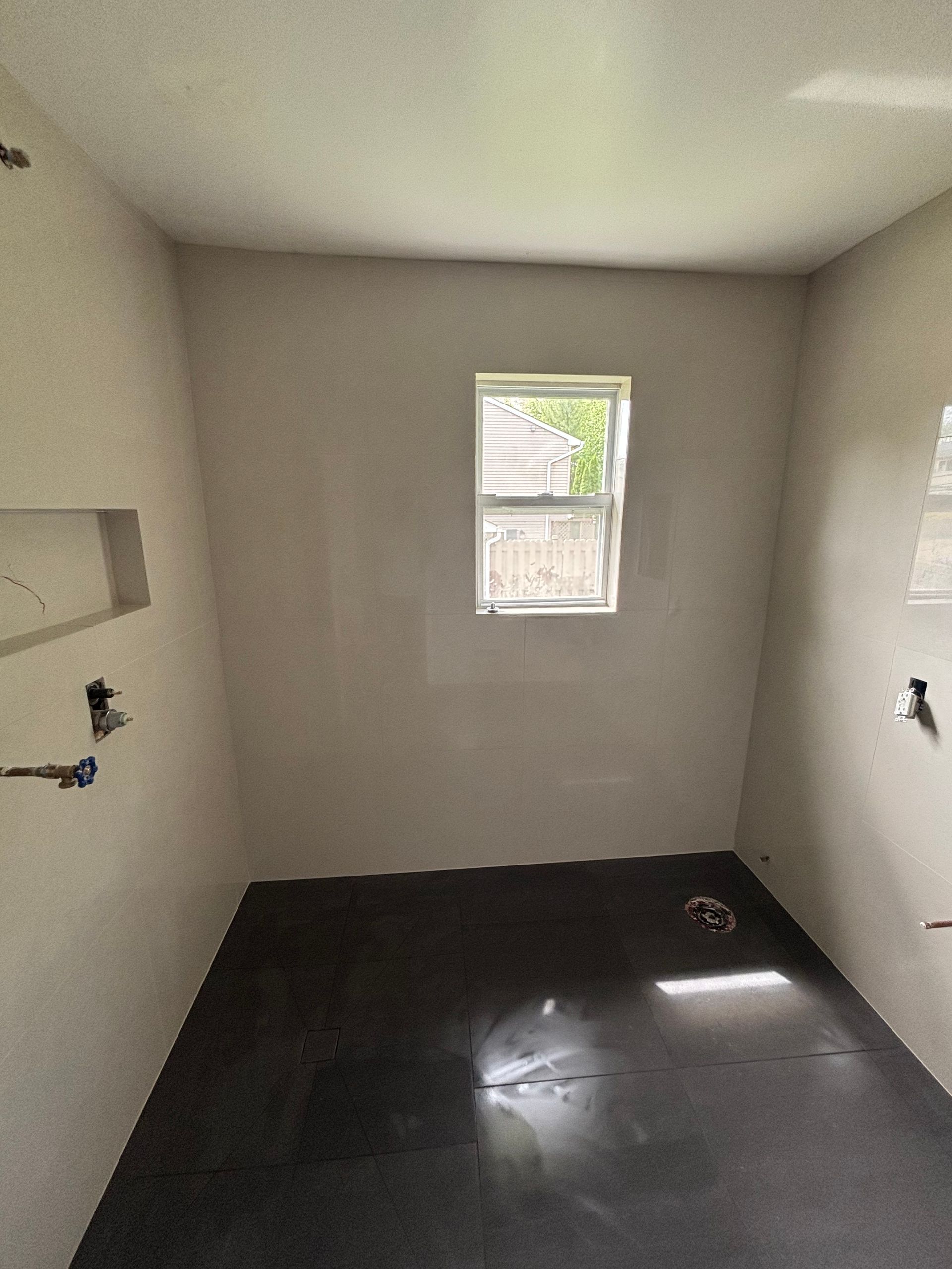 Empty bathroom, recently plastered walls, small window, dark floor.