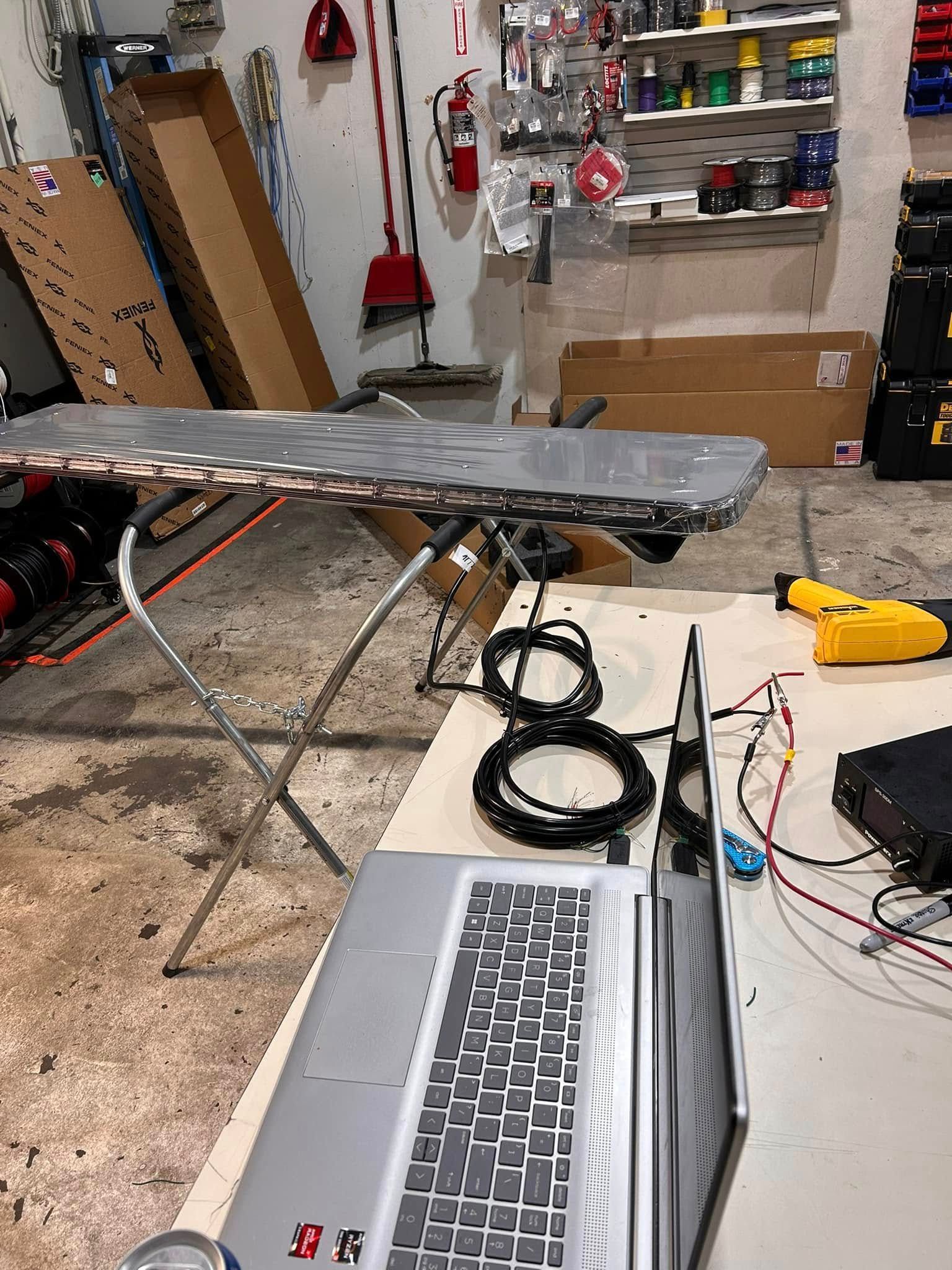 Laptop, wires, and equipment on a table, with a car part on a stand in a garage.