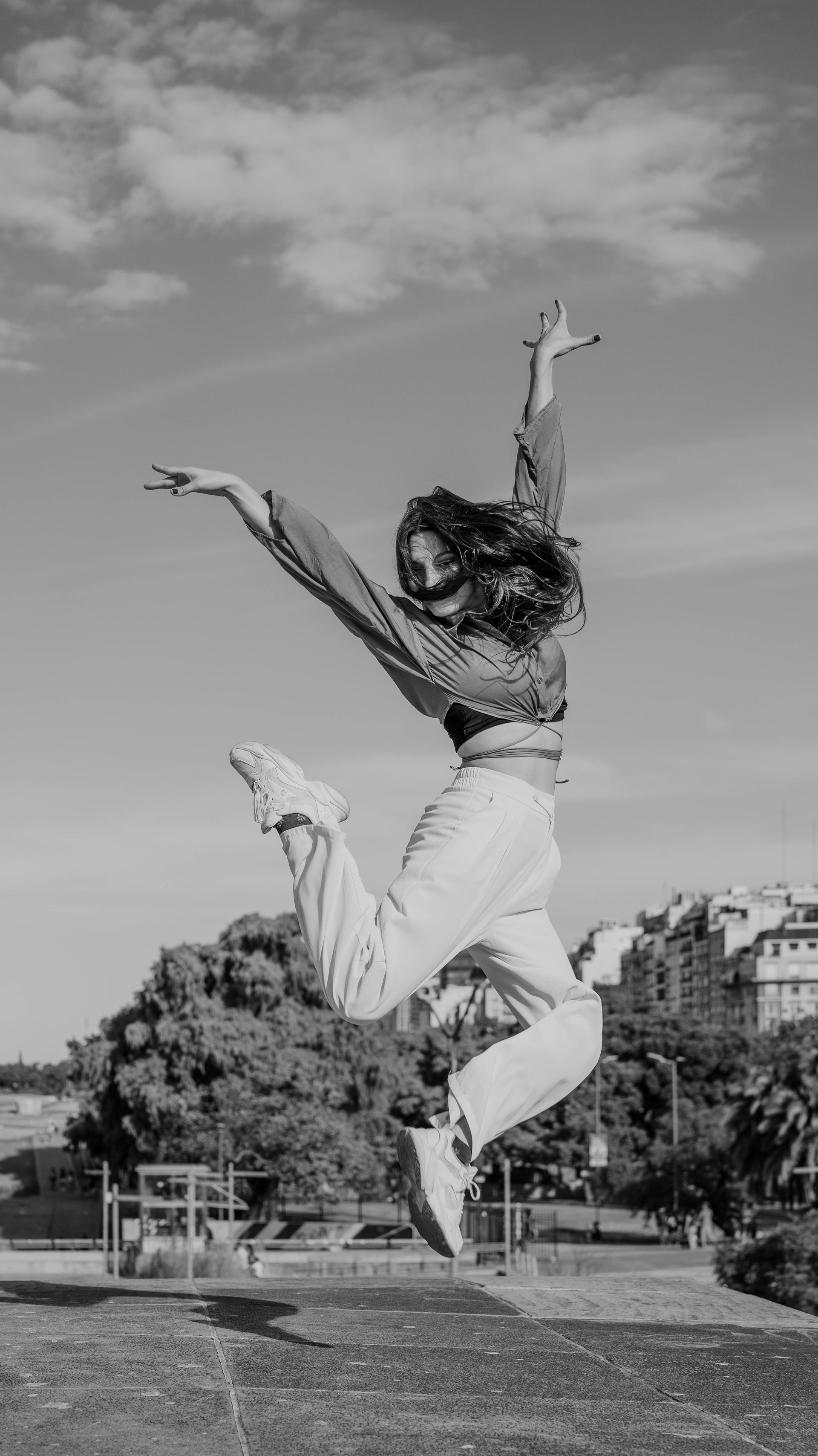 Une personne en pantalon blanc saute très haut, les bras levés, dans un parc sous un ciel nuageux.