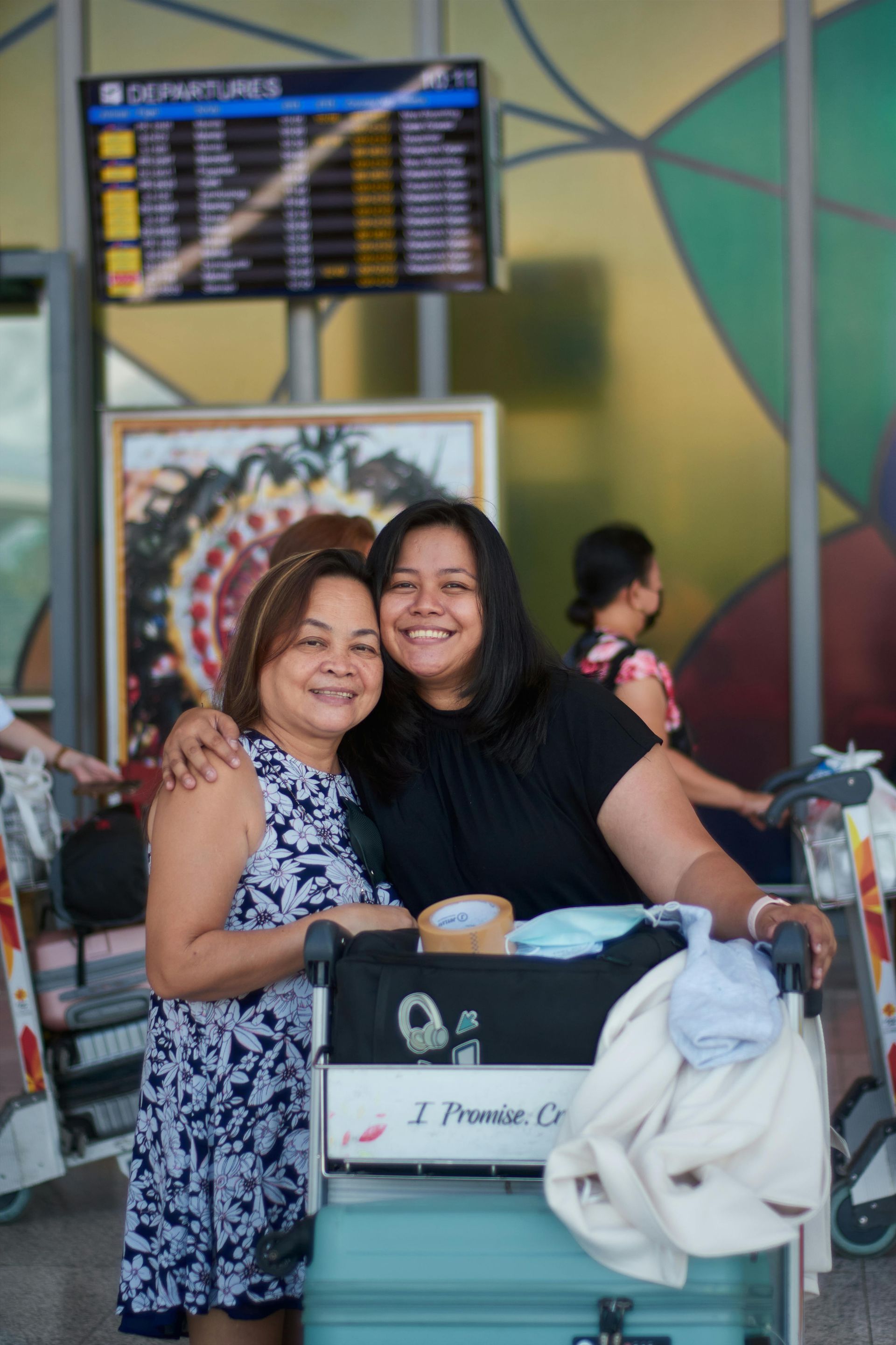 Two smiling women embrace at an airport, luggage cart in front of them. Departure board and colorful wall in the background.