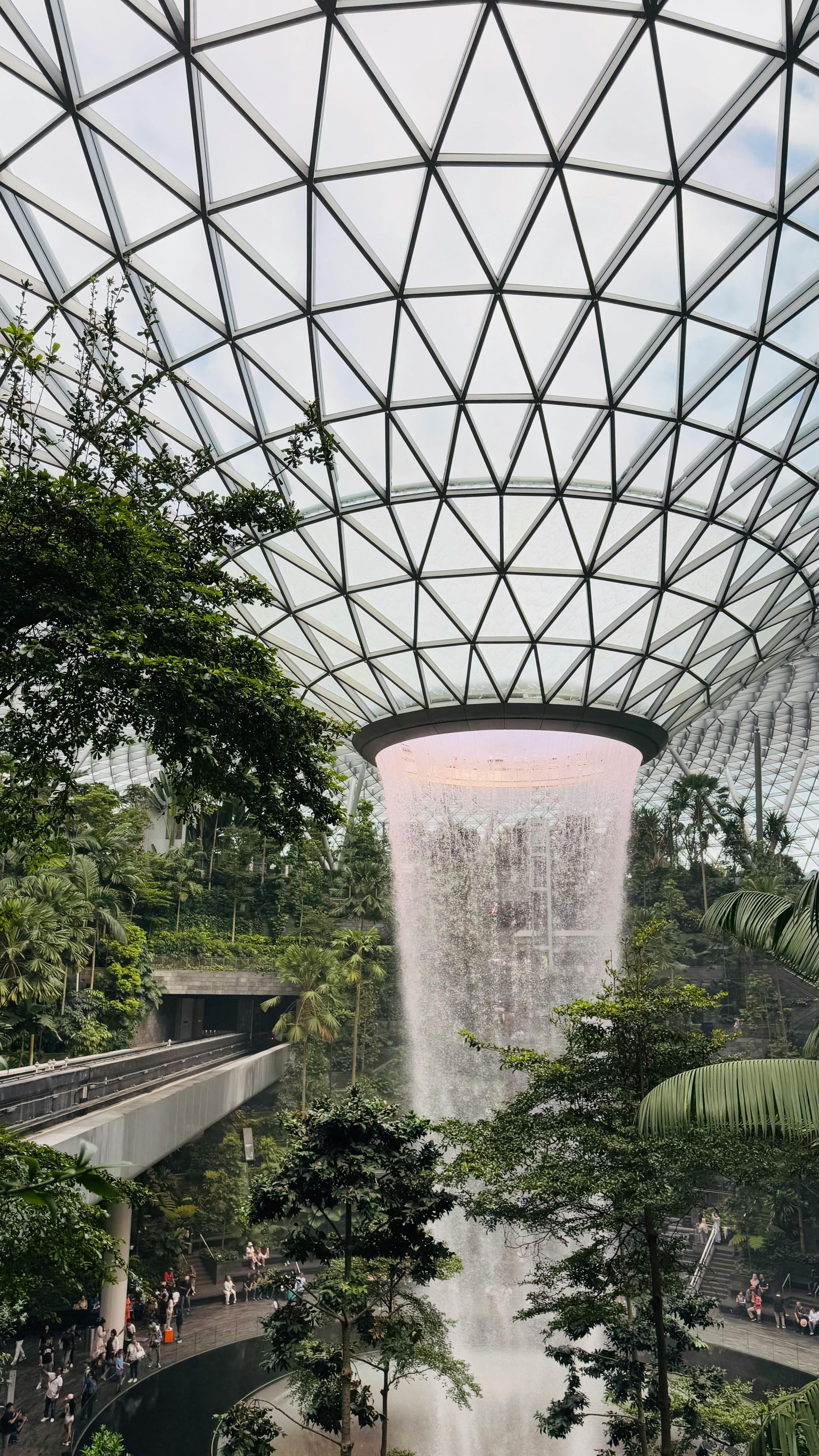 Indoor waterfall cascading from glass dome roof; lush greenery below. Jewel Changi Airport, Singapore.