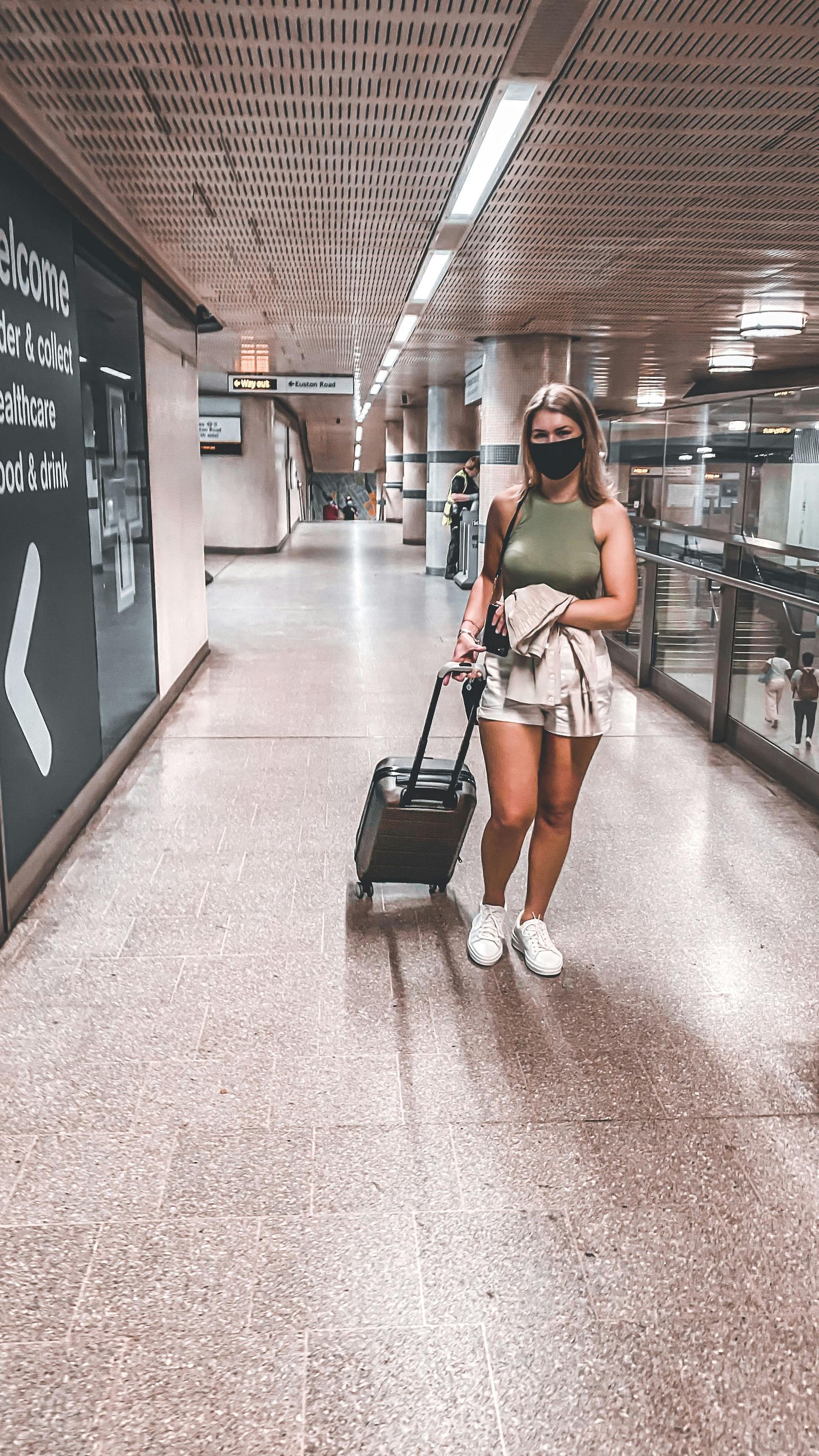 Woman in mask, tank top, shorts, and sneakers rolls luggage through an empty, long subway corridor.