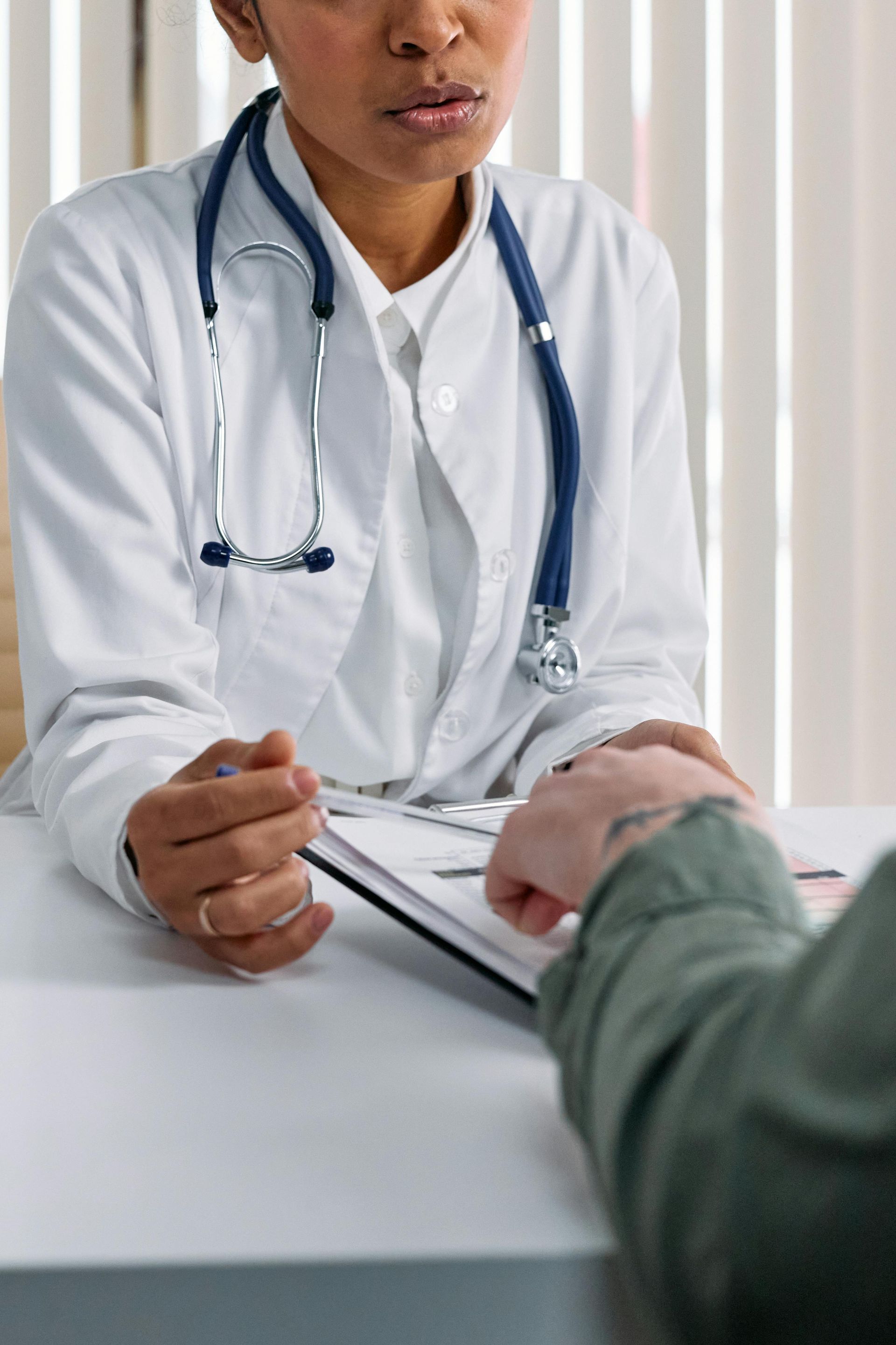 Doctor in a white coat with stethoscope consulting a patient at a desk.