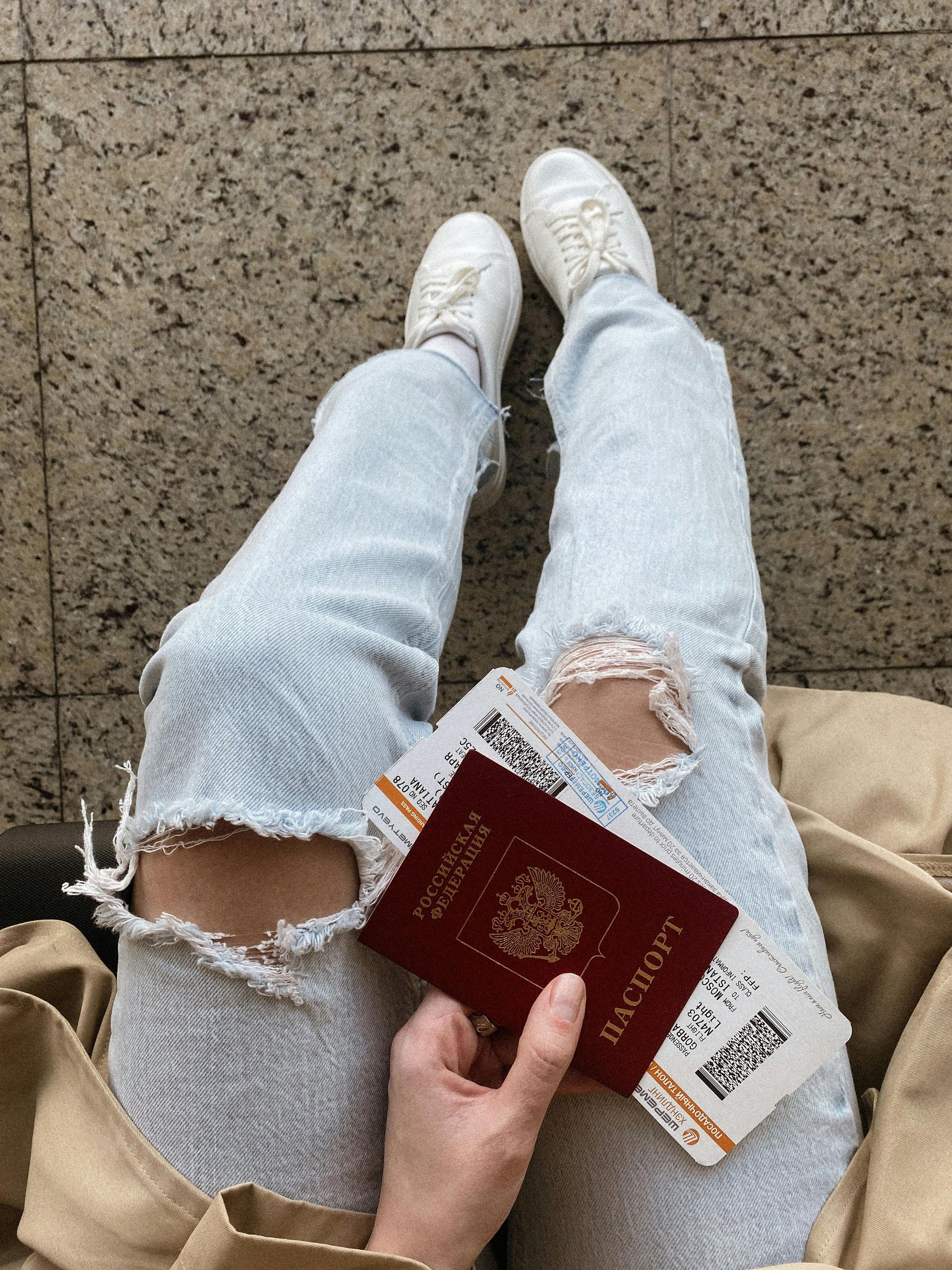 Person holding a Russian passport and boarding passes, wearing ripped light-wash jeans and white sneakers.