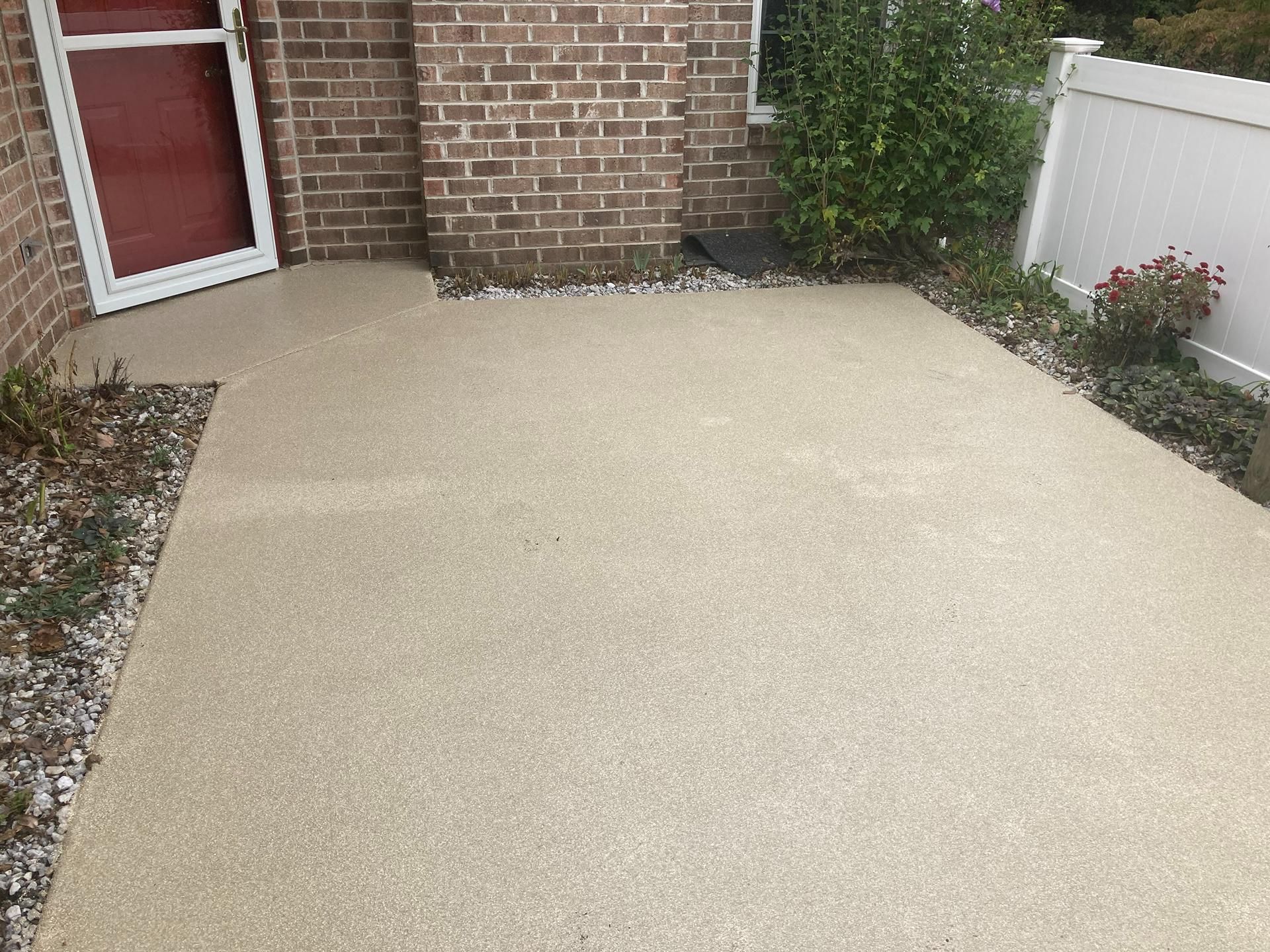 Beige concrete patio beside a brick house, with a red door and white fence.