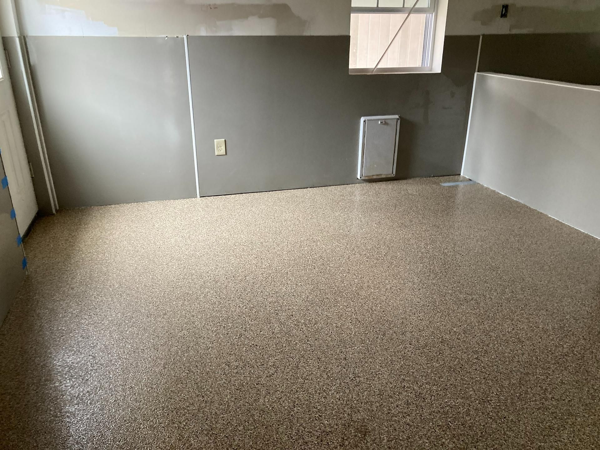 Empty tiled room with gray walls and a speckled floor, viewed toward a doorway and utility panel.