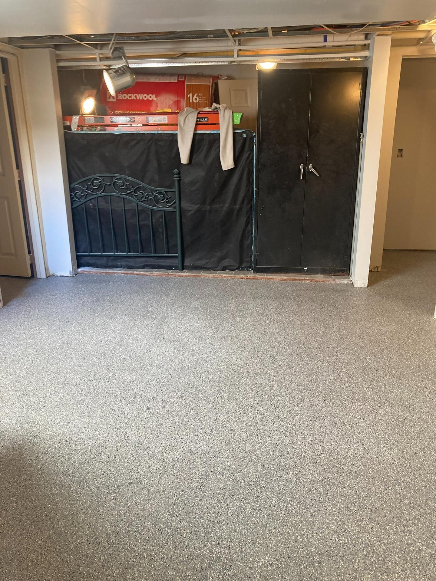 Small basement room with a black gate, hanging clothes, and a gray carpeted floor under exposed pipes.
