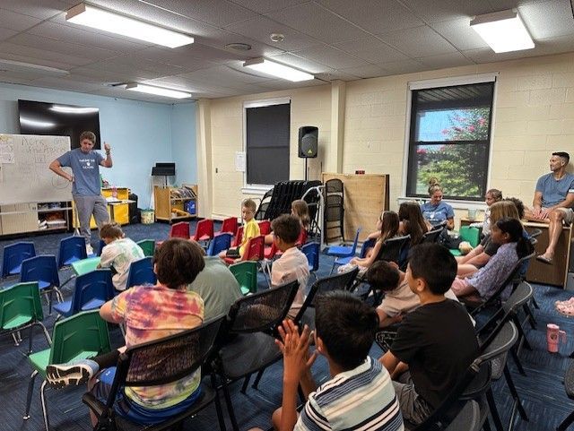 A man in a room with children, giving a presentation. Chairs are set up.