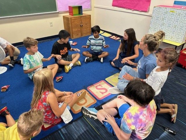 Children and teacher sit in a circle on a blue rug, playing a game with letter tiles in a classroom.